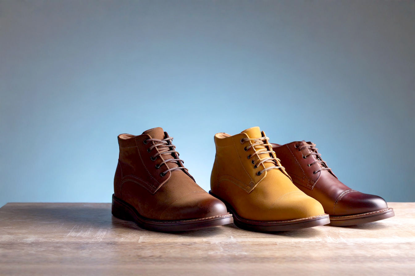 Professional photograph featuring three distinct pairs of men's chukka boots arranged in ascending formality order: a light tan suede desert boot with crepe sole, a medium brown leather chukka with subtle burnishing, and a polished dark brown calfskin chukka. The boots are photographed from a side angle on a neutral gray backdrop with soft, directional lighting highlighting the different textures and finishes of each leather type.