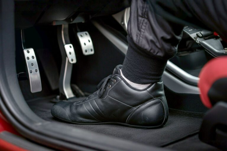 A close-up professional photograph of a driver's foot in a fireproof racing shoe pressing on a brake pedal inside a track car. The image should be taken from a side angle showing the thin sole profile of the racing shoe, with blurred motion in the background suggesting high-speed driving. The shoe should be clearly visible as a purpose-built racing shoe with visible FIA certification tags, contrasting against metal pedals that show signs of heat and use. The lighting should be dramatic with slight shadows to emphasize the texture and materials of the racing shoe.