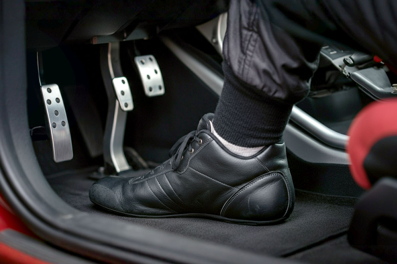 A close-up professional photograph of a driver's foot in a fireproof racing shoe pressing on a brake pedal inside a track car. The image should be taken from a side angle showing the thin sole profile of the racing shoe, with blurred motion in the background suggesting high-speed driving. The shoe should be clearly visible as a purpose-built racing shoe with visible FIA certification tags, contrasting against metal pedals that show signs of heat and use. The lighting should be dramatic with slight shadows to emphasize the texture and materials of the racing shoe.