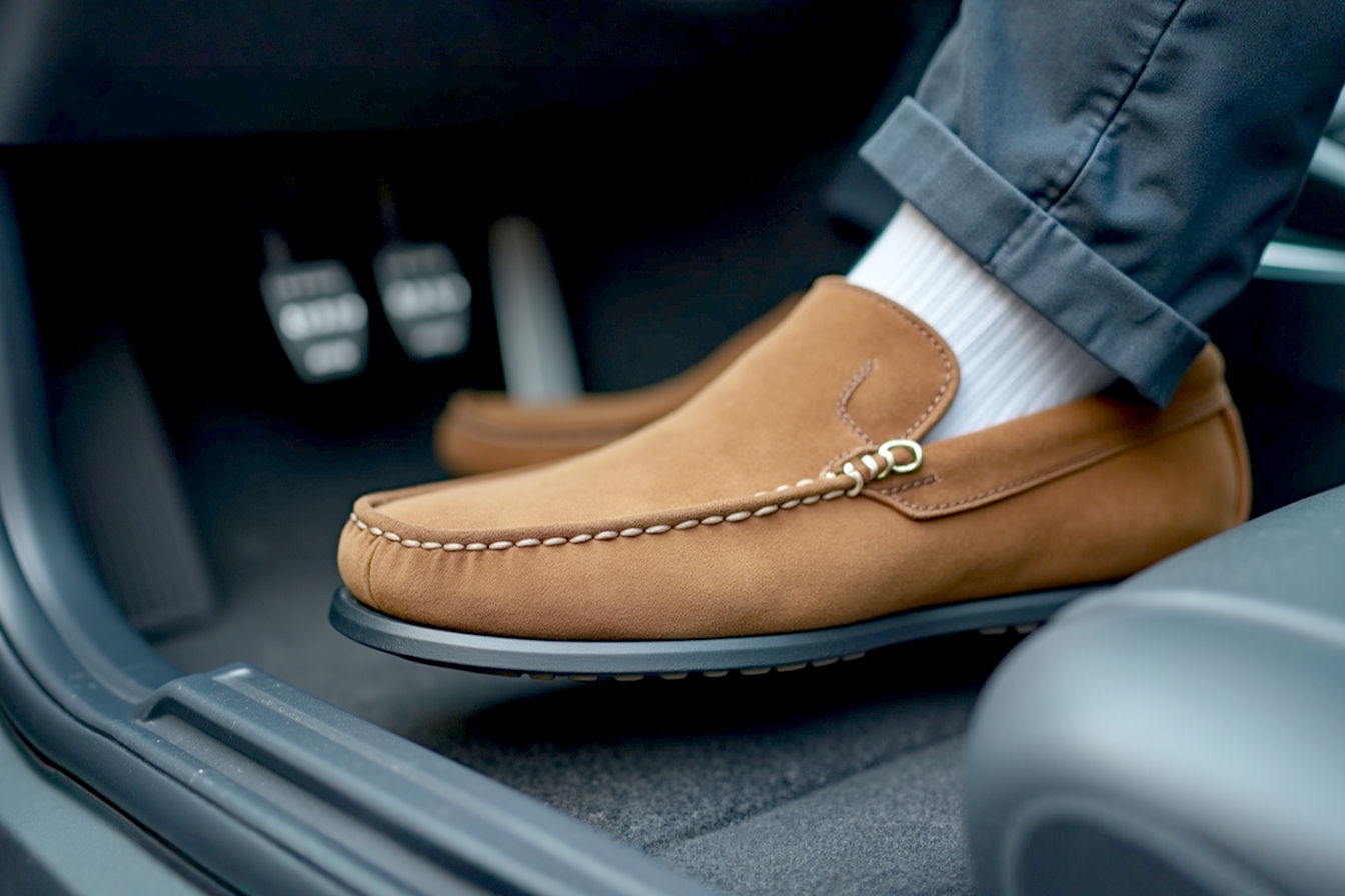 A high-quality, professional photograph showing a pair of stylish men's brown suede driving moccasins resting on the floor mat of a car, with the driver's foot lightly touching the accelerator pedal. The focus is on the shoe's sole and heel design, subtly highlighting the rubber pebbles and rounded heel. The background is slightly blurred, focusing attention on the shoe and pedal area. The lighting is natural, highlighting the texture of the suede and rubber.