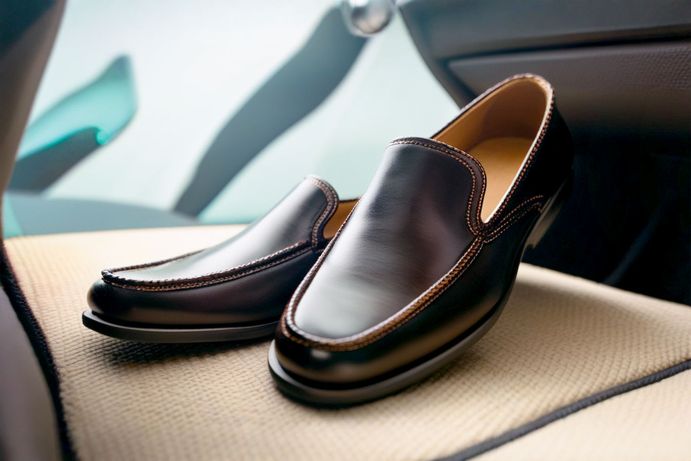 Professional, high-angle photograph showing a pair of dark brown leather driving slippers with a distinctive nubbed rubber sole, positioned on a clean car floor mat near the pedals of a luxury vehicle. The focus is sharp on the shoes and pedals, with the car interior slightly blurred in the background, conveying a sense of luxury and function.