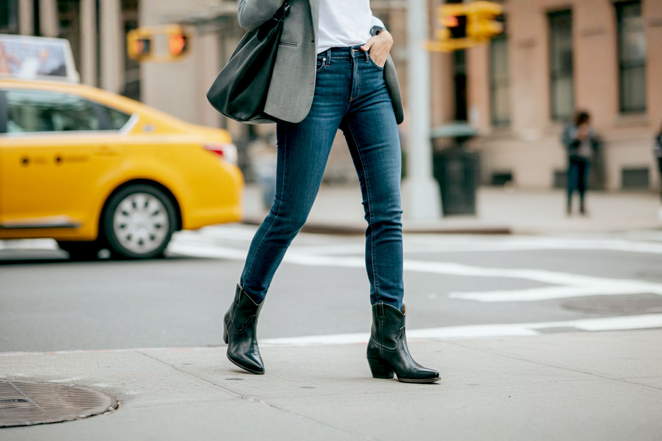 Professional street style photography featuring a stylish woman walking on a city sidewalk. She is wearing dark wash skinny jeans tucked into black leather short cowgirl boots with a moderate Cuban heel and subtle stitching. She has a tailored grey blazer over a simple white t-shirt and is carrying a structured black leather tote bag. The background shows blurred city buildings and a yellow taxi cab, emphasizing the urban setting. The image has a slightly desaturated, modern aesthetic.