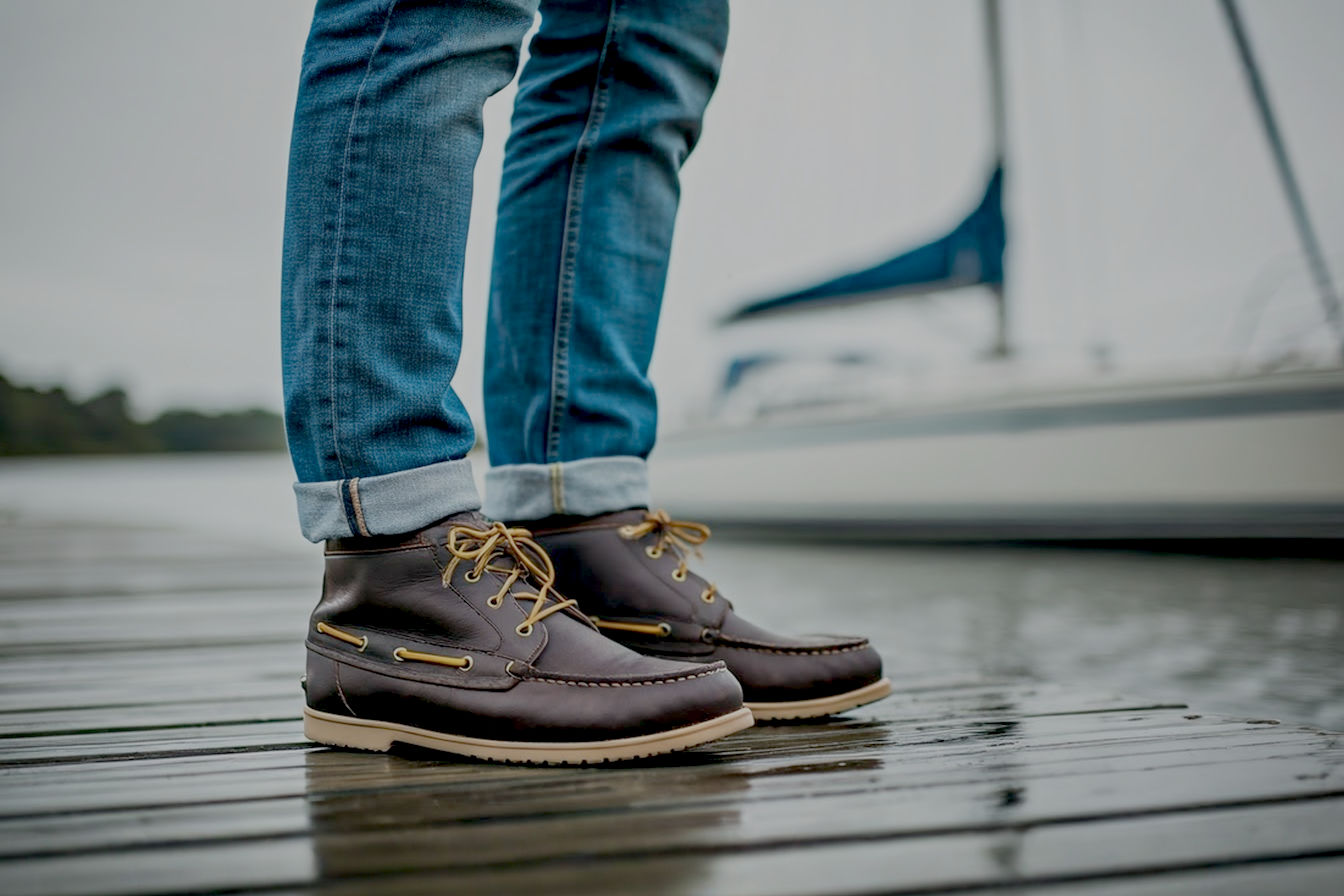 Professional photography style image of a man wearing stylish, waterproof leather boat boots standing on a wet wooden dock with a sailboat in the background. The boots are clearly visible and appear both rugged and fashionable.