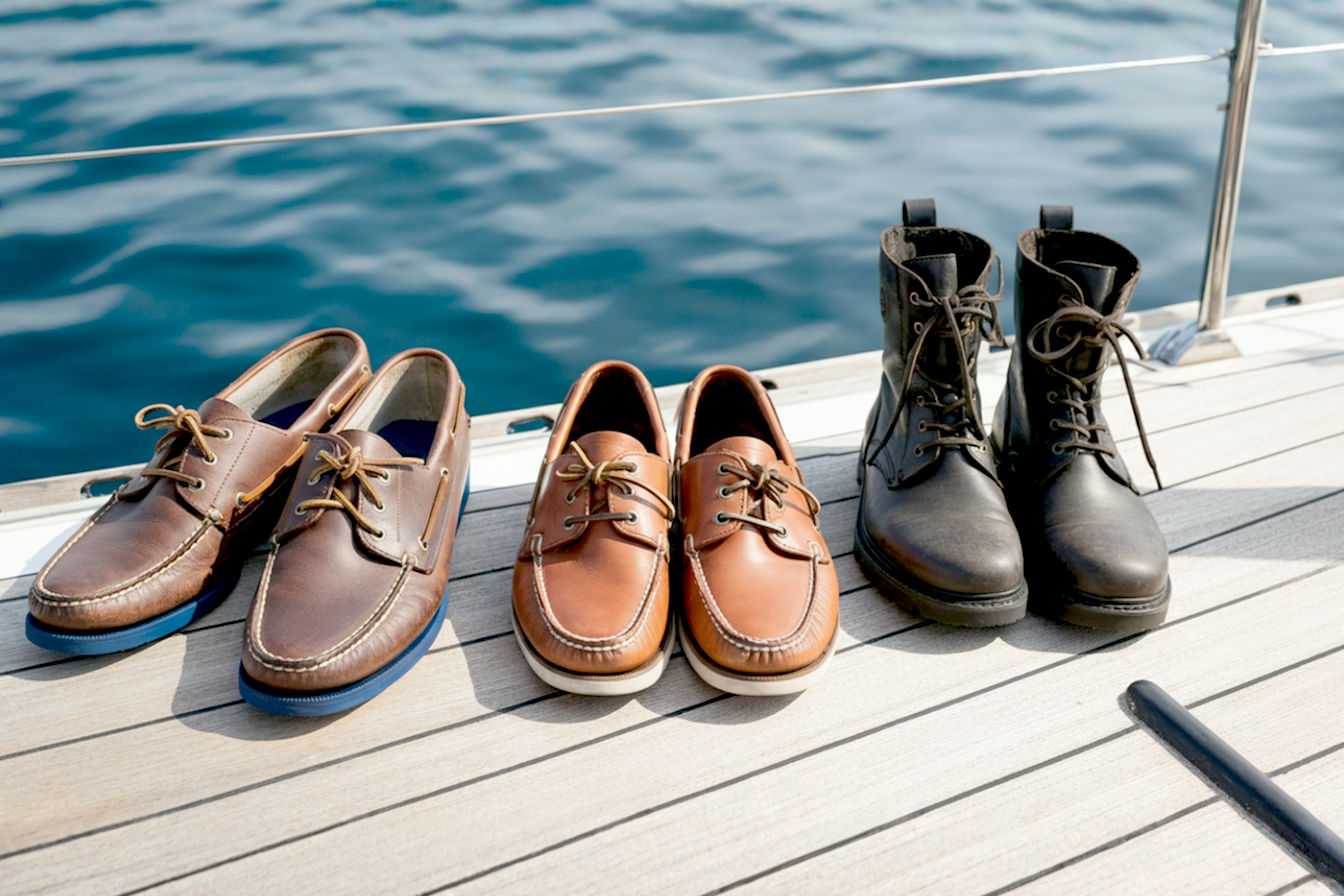 A professional overhead shot of a modern sailboat deck with three pairs of men's deck shoes arranged side-by-side. The shoes represent different styles: classic leather boat shoes, modern performance sailing shoes, and technical deck boots. The image captures sunlight gleaming on the deck and shoes, with blue water visible around the edges, conveying both the maritime setting and the importance of proper footwear choices.