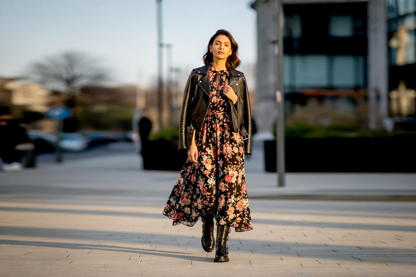 Professional, high-quality, full-body street style photograph of a stylish young woman (around 25-30 years old, diverse ethnicity) confidently walking on a chic urban street during the golden hour. She is wearing a flowing, romantic floral maxi dress (with a dark background and vibrant, medium-sized floral print) paired with classic black leather biker boots with buckles and a chunky sole. She also has a fitted black leather moto jacket casually draped over her shoulders. Her hair is effortlessly styled, and she has a subtle, confident expression. The lighting is soft and warm, casting long shadows. The background is slightly blurred (bokeh effect) but shows hints of modern city architecture. The photo should have a fashion magazine editorial quality, with crisp details and rich colors. Lifelike, pro photography style.
