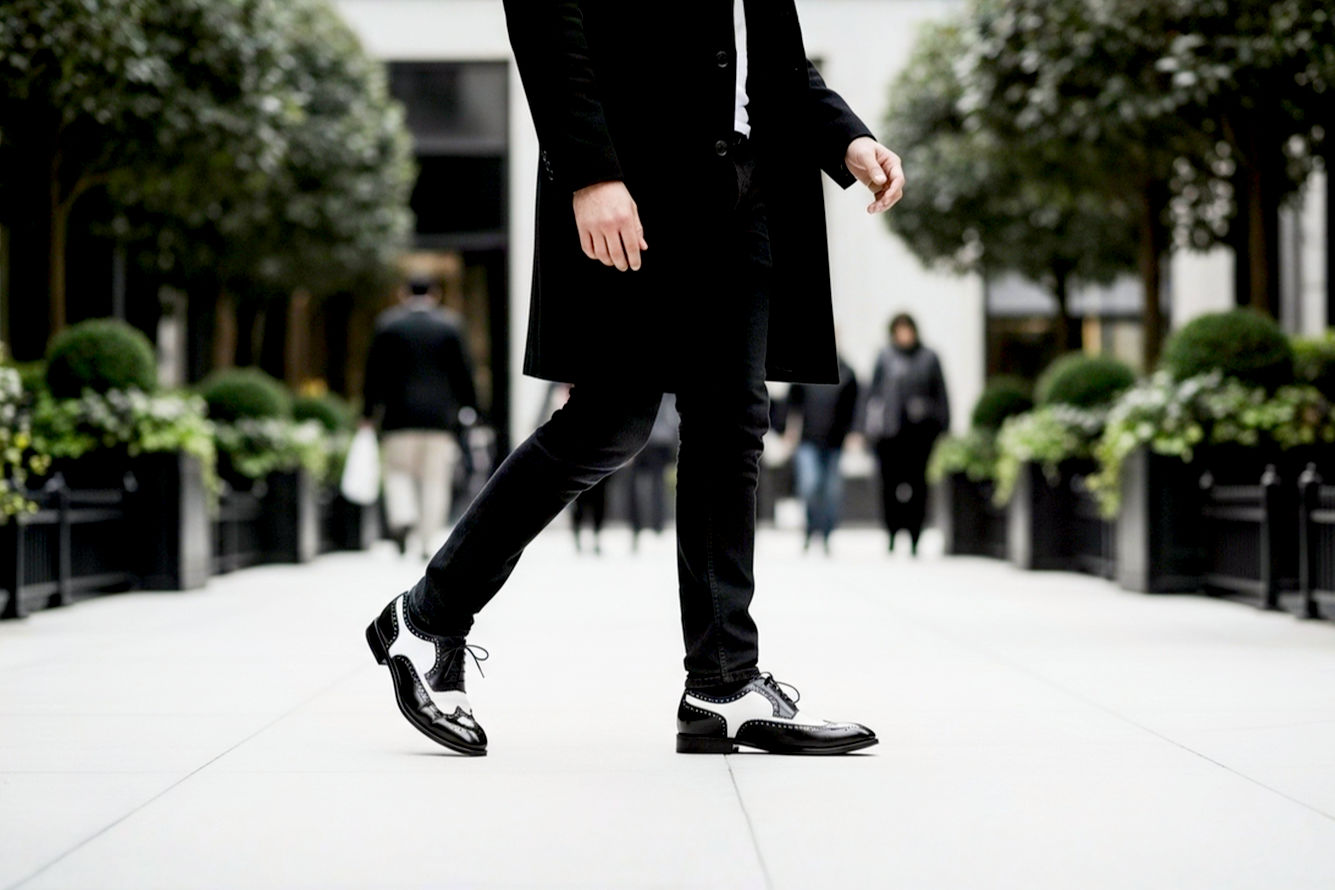 A professional, lifelike photograph of a stylish man, confidently walking on a clean, light-colored urban sidewalk. The focus should be on his feet, showcasing a pair of pristine, well-maintained black and white wingtip oxford shoes. The background should be slightly blurred, suggesting a sophisticated setting like an upscale city street or a garden party. The lighting should be soft and natural, highlighting the leather's texture and the sharp contrast of the shoe colors. The overall mood is confident, elegant, and timeless. No brands or logos visible.