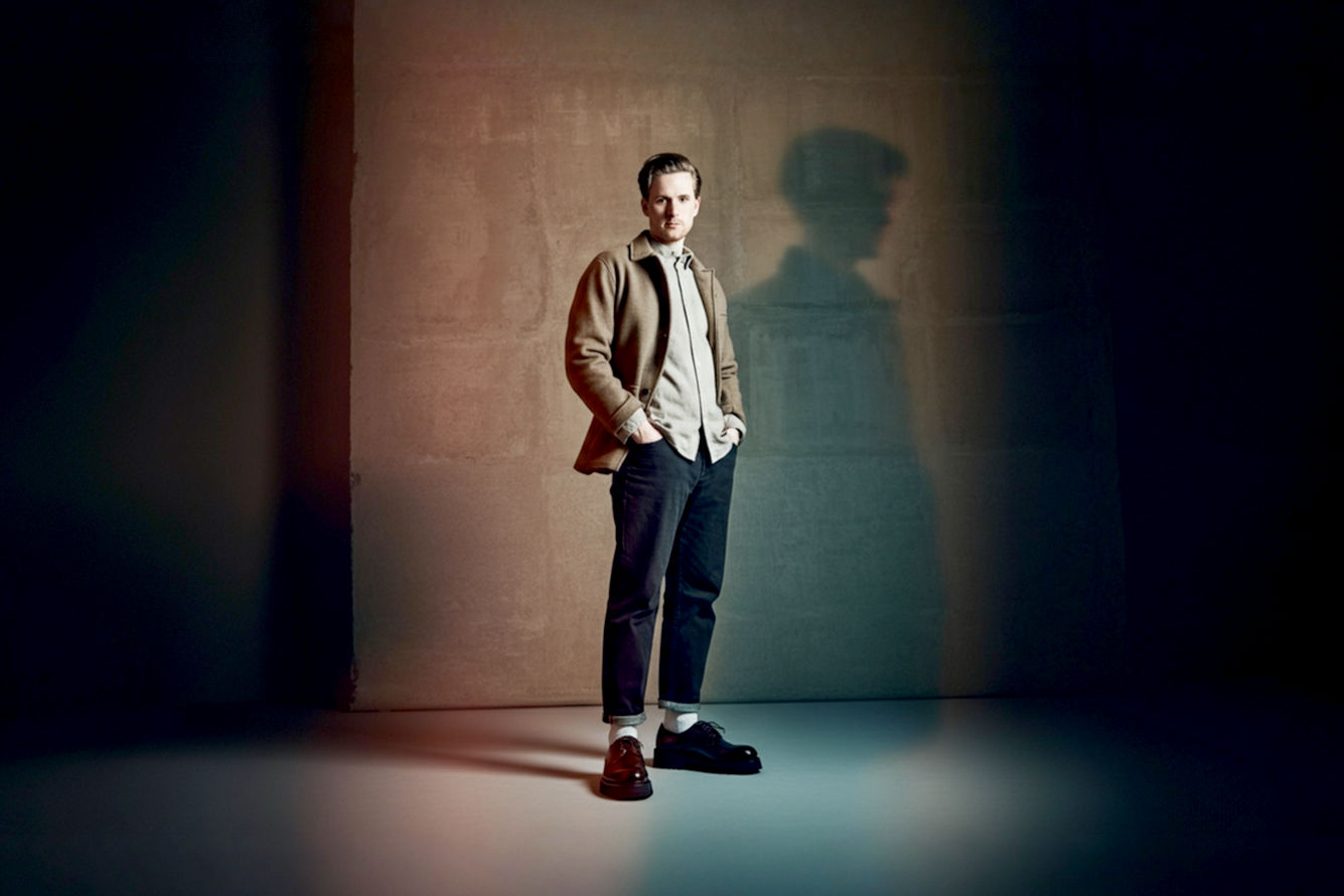 Professional, lifelike studio photography of a man (late 20s to early 30s) wearing stylish, dark brown chunky Derby shoes, standing with confidence. The man is dressed in smart-casual attire, possibly dark wash relaxed-fit jeans or tailored charcoal trousers, and a crisp, light-colored button-down shirt or a fine-gauge knit sweater. Focus on the shoes prominently in the foreground, showcasing their robust sole and quality leather texture. The background is a clean, minimalist studio setting with soft, diffused lighting, creating a sophisticated and modern atmosphere. The image should convey versatility, modern elegance, and a grounded, confident aesthetic, suitable for a men's fashion article.