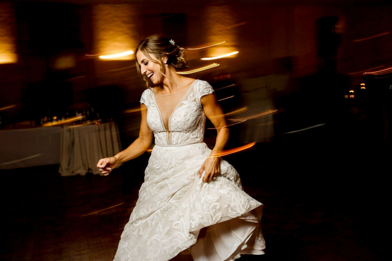A joyful bride with a radiant smile, seen from the waist down, dancing effortlessly at her wedding reception. Her elegant, low-heel bridal shoes are subtly visible. The lighting is warm and celebratory, capturing motion and happiness. The background is softly blurred, hinting at a lively dance floor or beautiful wedding venue. Lifelike pro photography style, focus on genuine emotion and graceful movement.