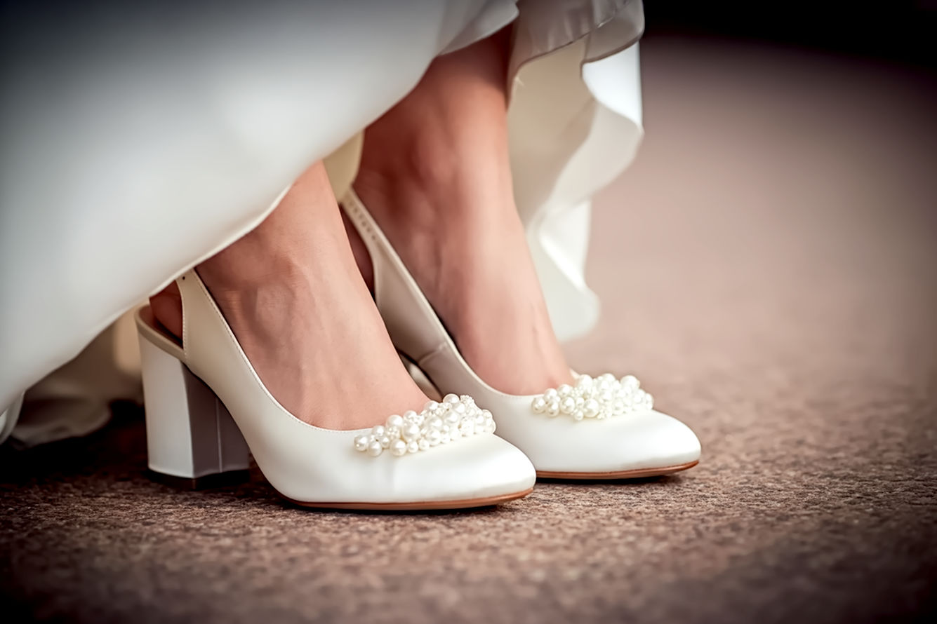 Lifelike pro photography of a close-up of a bride's feet wearing elegant ivory or white block heels adorned with delicate, shimmering pearls. The bride is standing on a beautiful, slightly textured surface, possibly a subtly blurred garden path or a polished ballroom floor. The lighting is soft and ethereal, highlighting the pearls' luster and the shoes' texture. The bride's wedding dress hem is visible, gracefully flowing around the shoes. The image should convey comfort, elegance, and sophistication, with a shallow depth of field that emphasizes the shoes. Shot with a prime lens for beautiful bokeh. Warm, inviting tones.
