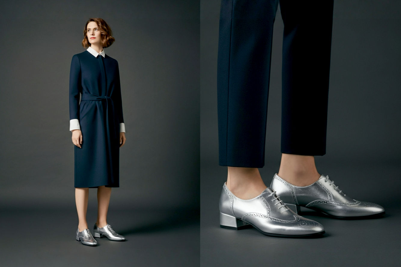 Professional photography style, high-resolution, lifelike image. A stylish woman, mid-30s, standing confidently. She is wearing a modern, well-tailored outfit that is balanced and chic, with a strong focus on her silver low block heels. Her outfit features a deep navy midi dress made of a matte, rich fabric (like crepe or wool blend) or perfectly tailored, ankle-length black trousers with a crisp white blouse. The silver heels are prominent, reflecting subtle light, showcasing their block heel and low height. The background is clean, sophisticated, and slightly blurred, perhaps an urban chic setting or a minimalist interior, ensuring the focus remains on the fashion. The lighting is soft and flattering, highlighting the metallic sheen of the shoes without harsh glares. The overall mood is elegant, modern, and confident.