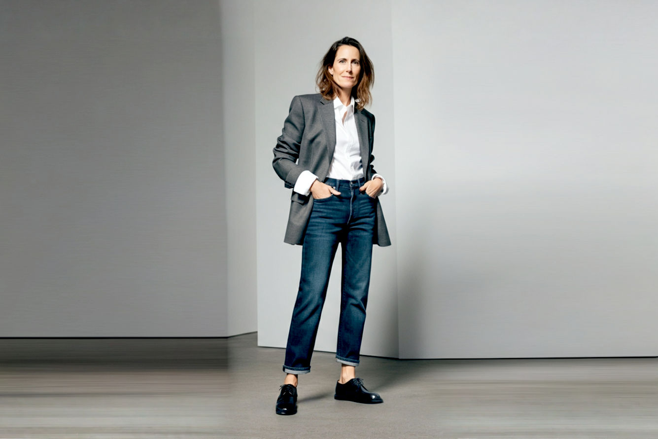 Professional photography of a woman (mid-30s) standing confidently. She is wearing impeccably styled black leather Derby shoes with a chunky sole, paired with tailored, dark-wash, straight-leg jeans that are slightly cropped to showcase the shoes. Her upper body features a crisp white button-down shirt layered under an oversized yet structured charcoal blazer. Her pose is natural, with one hand casually in her pocket, projecting a sense of power and chic elegance. The background is a minimalist, modern urban setting (e.g., a blurred, clean street or architectural interior) that emphasizes her presence. Lighting is soft, diffused natural light that highlights the texture of the shoes and clothing. The image should convey sophistication, versatility, and quiet strength, with a focus on the footwear and overall stylish ensemble. High resolution, lifelike, pro photography style.