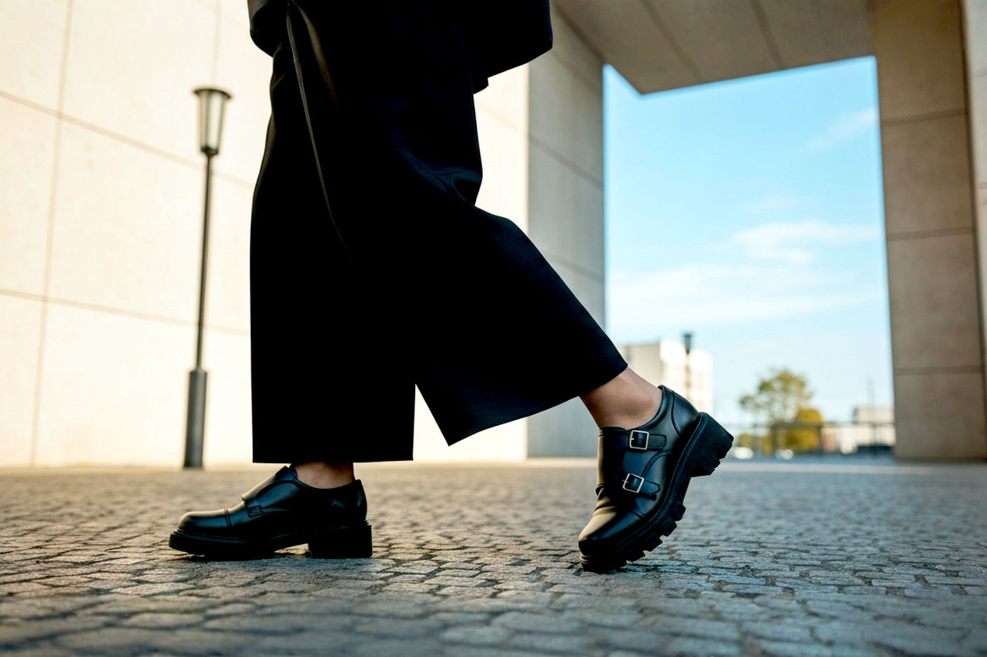 A stylish person, mid-stride, confidently showcasing chunky black double monk strap shoes. The focus is on the shoes and how they seamlessly integrate into a chic, balanced outfit. For a male model: A slim-fit, dark-colored suit or smart casual attire (e.g., tailored chinos and a blazer). For a female model: A flowing midi skirt/dress or wide-leg tailored trousers. The setting is an urban, modern environment (e.g., a cobblestone street, a minimalist architectural backdrop). Lighting is natural and bright, highlighting textures and details. Lifelike, professional photography style with a shallow depth of field, making the shoes pop.