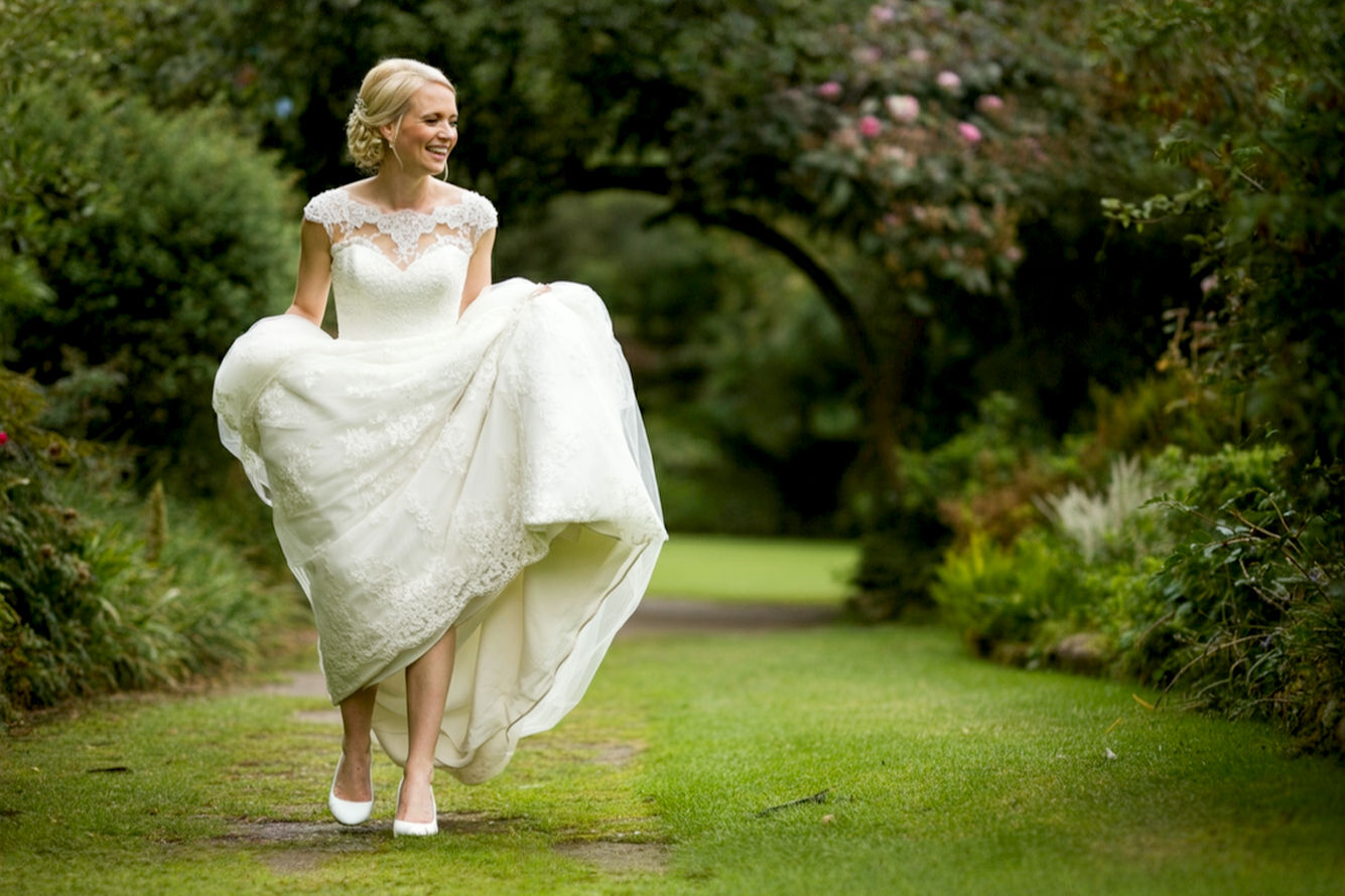 Lifelike professional photography of a modern bride confidently and joyfully walking in white kitten-heel bridal shoes on an outdoor wedding path, with a soft focus on her comfortable, elegant footwear. The bride is smiling, looking graceful and at ease, perhaps with a wedding dress flowing around her ankles. Emphasize the comfort and style of the shoes. Natural lighting, dreamy wedding aesthetic, high detail.