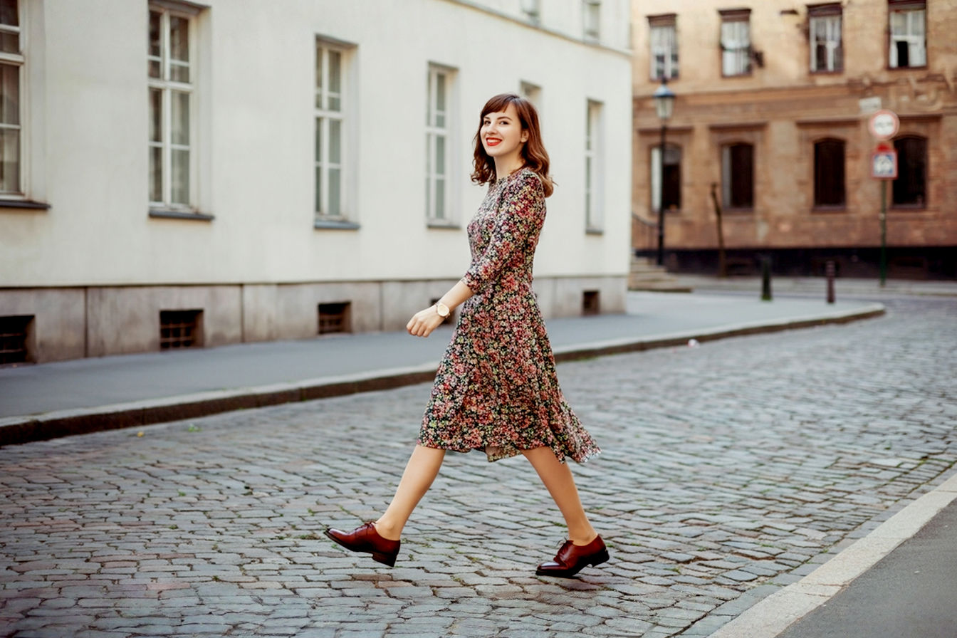 A stylish woman in her late 20s confidently walking down a cobblestone street, wearing a chic knee-length floral dress paired with classic leather brogues. The shot is a full body, slightly low-angle perspective, capturing her in motion with a natural smile. The background is softly blurred with urban architectural elements. Pro photography style, natural lighting, sharp focus on the subject, vibrant colors.