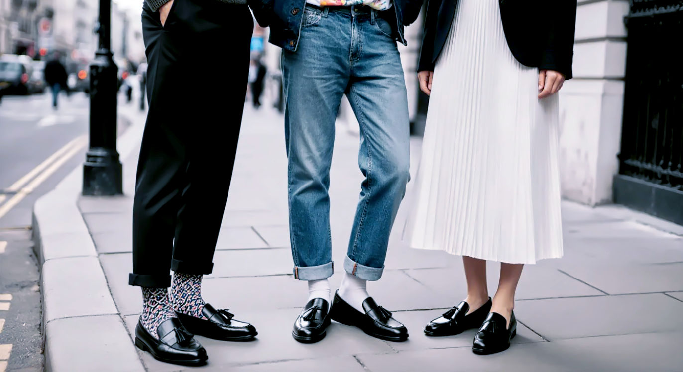 A dynamic, full-body street style photograph of three stylish individuals standing together on a London street corner, each showcasing a different way to wear the Adrian black tassel loafer.
Person on the left wears them in a "Classic" style with cropped tailored trousers, a knitted polo shirt, and visible patterned socks.
Person in the centre has a "Casual" look, pairing the loafers with turned-up selvedge denim, a vintage band t-shirt, and a chore jacket.
Person on the right showcases a "Modern Twist," wearing the loafers with a pleated midi skirt and an oversized blazer.
The photo is shot in the style of a high-end fashion blog, with natural afternoon light, capturing a candid, confident moment.